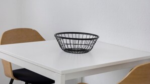 A white table with a black wire basket on it, wooden chairs, and a white wall.
