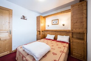 A bedroom with a wooden headboard, a bed with a patterned bedspread, a wall-mounted picture, and a wooden door. - Relaxing Duplex with Pool in the Mountains (Fontcouverte-la-Toussuire)