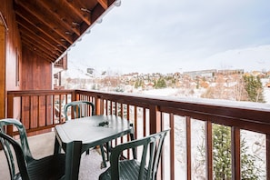 A wooden balcony with a table and chairs, overlooking a snowy village with houses and mountains. - Relaxing Duplex with Pool in the Mountains (Fontcouverte-la-Toussuire)