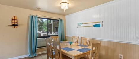 Un comedor con una mesa y sillas de madera, una araña, un ventanal con cortinas y una pala en la pared.
