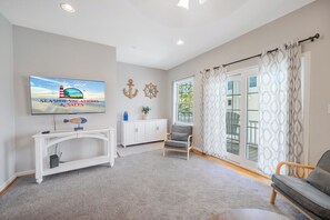 Una sala de estar con televisor, una consola blanca, un sillón gris, una silla de madera y un ventanal con cortinas estampadas.