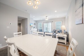 A modern dining area with a white table and chairs, a kitchen in the background, and a view of the outdoors through large windows. - Captain View Villa 3 by Seaside Vacations & Sales (Ocean City)