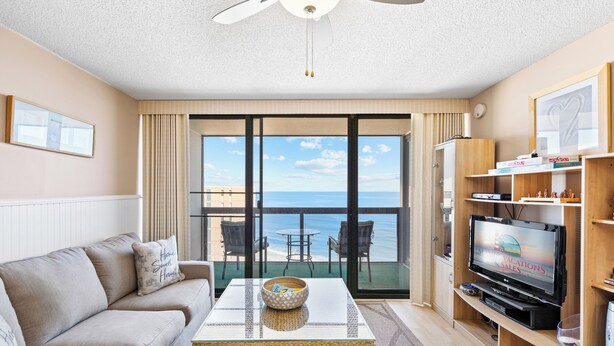 A living room with a sofa, coffee table, TV, and a balcony with a view of the ocean. - Ocean View condo w/ 3 pools by Seaside Vacations & Sales (Ocean City)