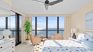 Une chambre d’hôtel avec un grand lit, un ventilateur de plafond, une vue sur l’océan, une plante et un bureau avec une lampe.