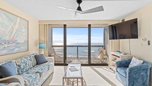 Un salon spacieux doté d’une grande fenêtre offrant une vue sur la mer, d’un ventilateur de plafond et d’un téléviseur à écran plat fixé au mur.