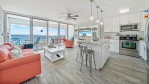 Eine moderne Küche mit weißen Schränken, einer Mikrowelle und einem Backofen. Es gibt eine Bar mit Hockern und einem Fernseher, der eine Strandansicht zeigt.