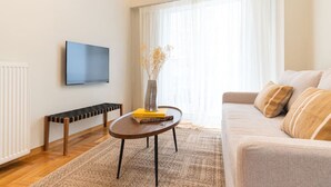 A modern living room with a sofa, a coffee table, a TV mounted on the wall, and a window with sheer curtains. - The MP10 Residence - Kolonaki (Athens)