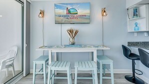 A modern kitchen with a seaside-themed TV, a wooden table with chairs, and a bar area with a blue bottle. - Sea Gate 202 by Seaside Vacations & Sales (Ocean City)