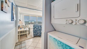 A laundry room with a white washer and dryer, a view of a living area with a sofa and a chair, and a welcome mat at the entrance. - Ocean view condo Seaside Vacations & Sales (Ocean City)