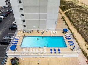 An aerial view of a hotel with a swimming pool surrounded by sun loungers, a parking lot, and a sandy area with umbrellas. - Stunning oceanfront condo by Seaside Vacations & Sales (Ocean City)