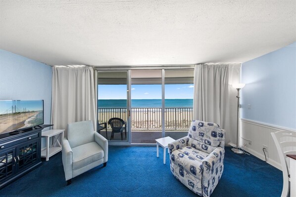 A room with a blue carpet, a sofa, a chair, a TV, and a balcony with a view of the beach and ocean. - Stunning oceanfront condo by Seaside Vacations & Sales (Ocean City)