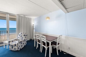 A room with a balcony, a dining table with chairs, a patterned sofa, and a view of the beach. - Stunning oceanfront condo by Seaside Vacations & Sales (Ocean City)
