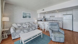 Un espace de vie compact comprenant un canapé, un fauteuil, une petite table, un coin cuisine avec des meubles blancs et des appareils électroménagers, et un tableau encadré accroché au mur.