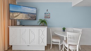 Habitación con televisor sobre un mueble bajo, una pequeña mesa con sillas, una planta en maceta y un cartel en la pared.