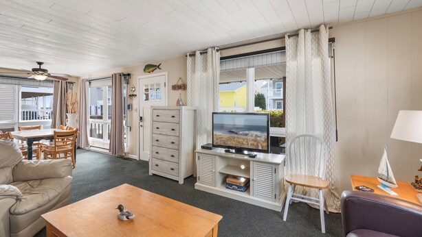 A living room with a television, wooden furniture, a dining area, and a view of the sea through large windows. - Seas the Day 1 by Seaside Vacations & Sales (Ocean City)