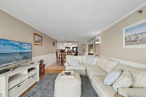 Una sala de estar con un televisor de pantalla plana grande, un sofá a rayas, una mesa de centro y un área de comedor al fondo.