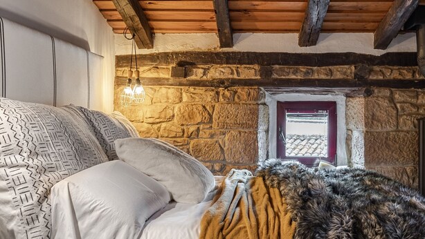 A cozy bedroom with a bed, a stone wall, a wooden ceiling, and a small window.