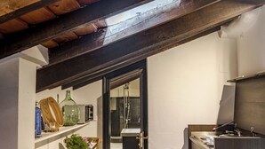 A narrow kitchen with dark wooden beams, a white table with a chair, and a small potted plant.