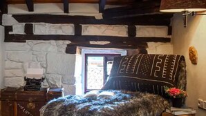 A cozy room with a stone wall, wooden beams, a fur-covered bed, a lamp, and a small table with a book.