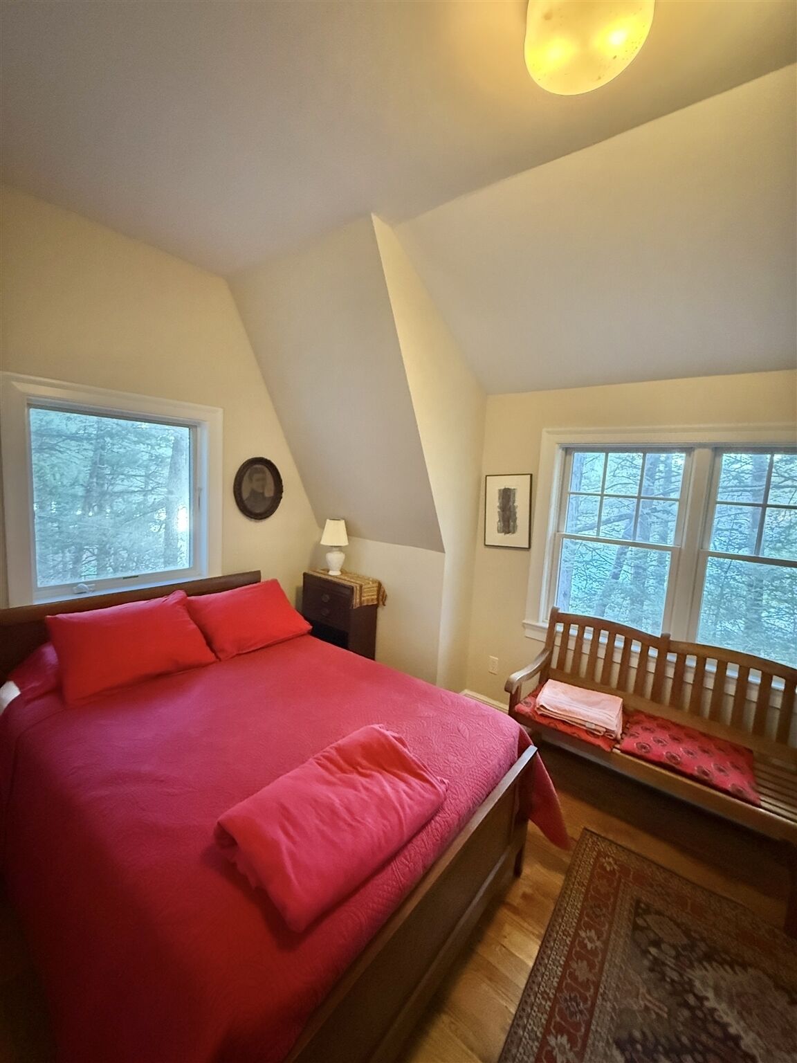 A bedroom with a bed, a nightstand, a lamp, a bench, and two windows with a view of trees.