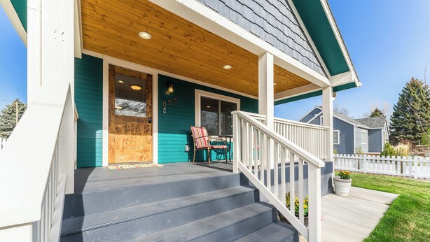A front porch with a wooden deck, a covered entryway, and a small outdoor seating area with a chair and table. - Kanon’s Kottage | Downtown Retreat Near RMNP (Loveland)
