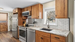 A modern kitchen with wooden cabinets, stainless steel appliances, a granite countertop, and a window with a view of trees. - Kanon’s Kottage | Downtown Retreat Near RMNP (Loveland)