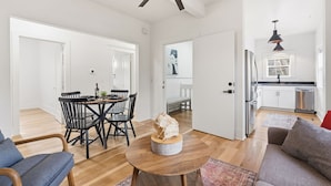 Ein geräumiger Wohnbereich mit Esstisch, Stühlen, einem runden Couchtisch und einer Couch. Es gibt eine Küchenzeile mit Kühlschrank, Herd und Spüle. Der Raum verfügt über Holzdielenböden und weiße Wände.