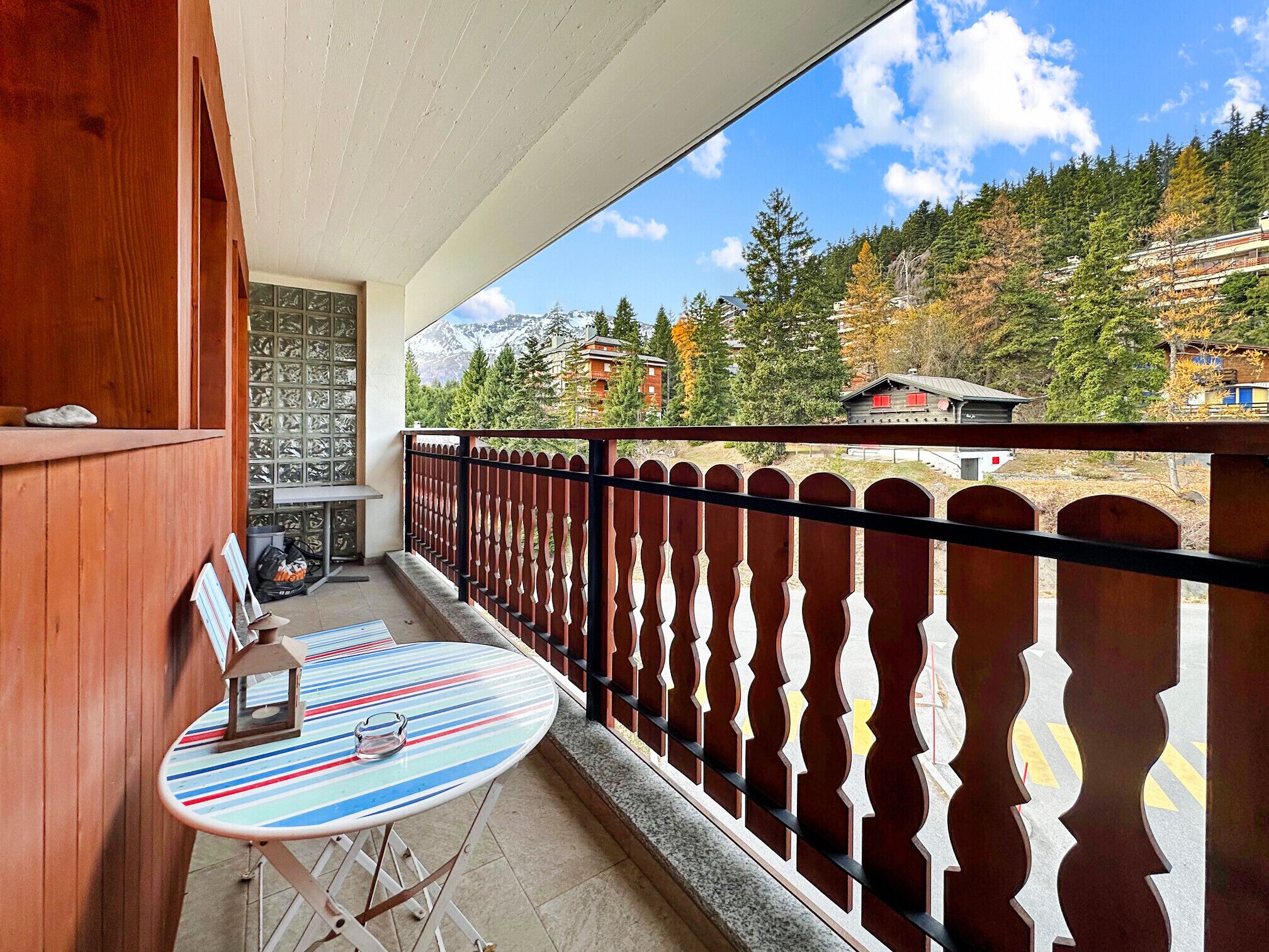Een balkon met een gestreepte tafel en stoelen, een houten leuning en uitzicht op een bergachtig landschap met gebouwen.
