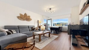 Ein modernes Wohnzimmer mit grauem Sofa, Couchtisch aus Holz und einem Essbereich mit Blick auf eine gebirgige Landschaft.