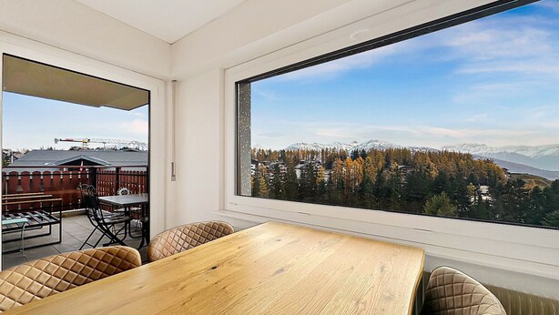 Ein modernes Zimmer mit einem großen Holzespeisetisch und gepolsterten Stühlen. Ein großes Fenster bietet Blick auf eine gebirgige Landschaft mit Bäumen und Gebäuden in der Ferne.