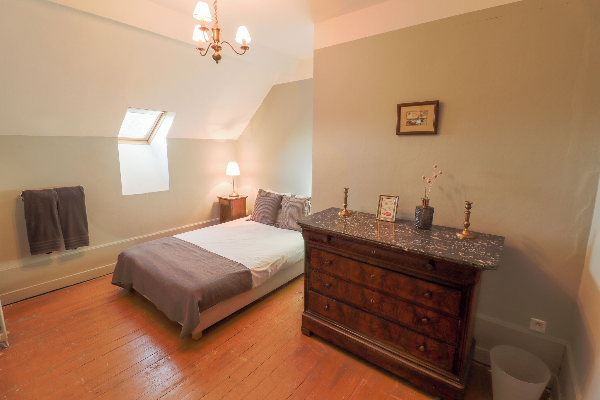 Une chambre à coucher avec un lit, une commode en bois, une table de chevet, une lampe et une petite fenêtre.