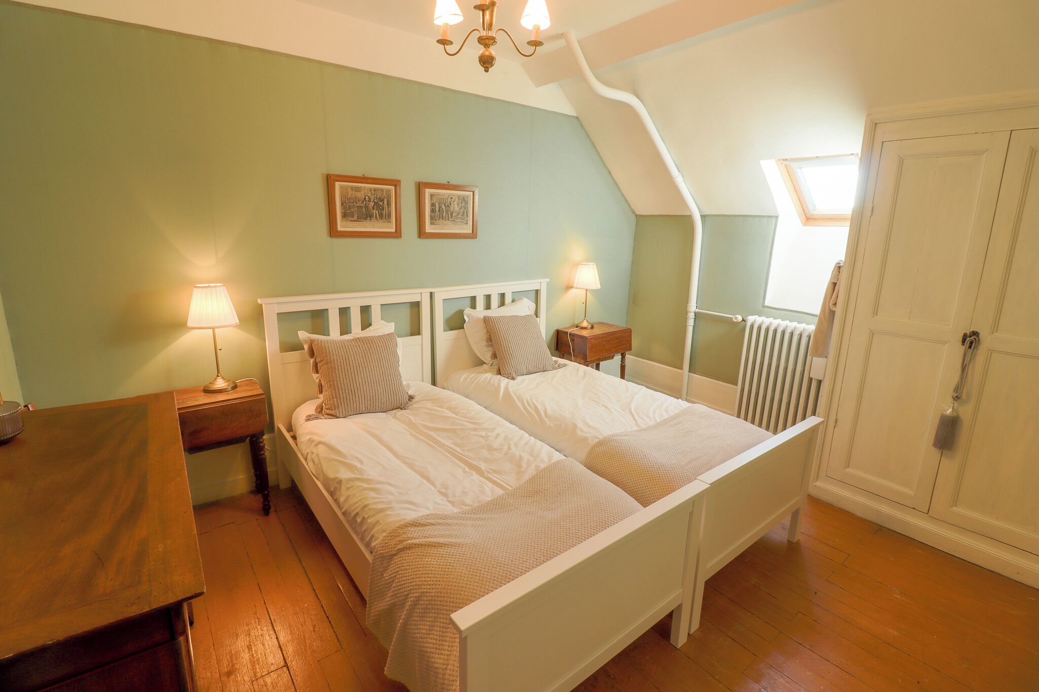 A bedroom with two beds, a wooden nightstand, a desk, a radiator, and a door.