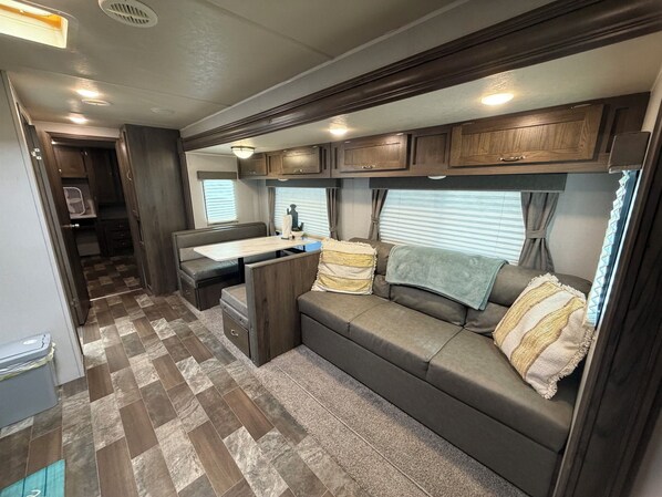 A compact RV interior with a kitchenette, a dining area, and a seating area with a sofa and cushions.