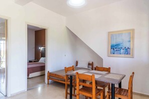 A room with a dining table and chairs, a bed with a pink bedspread, and a painting on the wall. - Pink House Socrates Corfu Beachside Apartments (Gardenos)