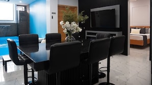 A modern hotel room with a dining area featuring a black table and chairs, a large flat-screen TV, and a small pool area.