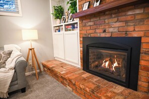 Ein gemütliches Wohnzimmer mit Kamin, einem grauen Sofa, einer Lampe und einer weißen Regaleinheit mit Büchern und Dekoration.