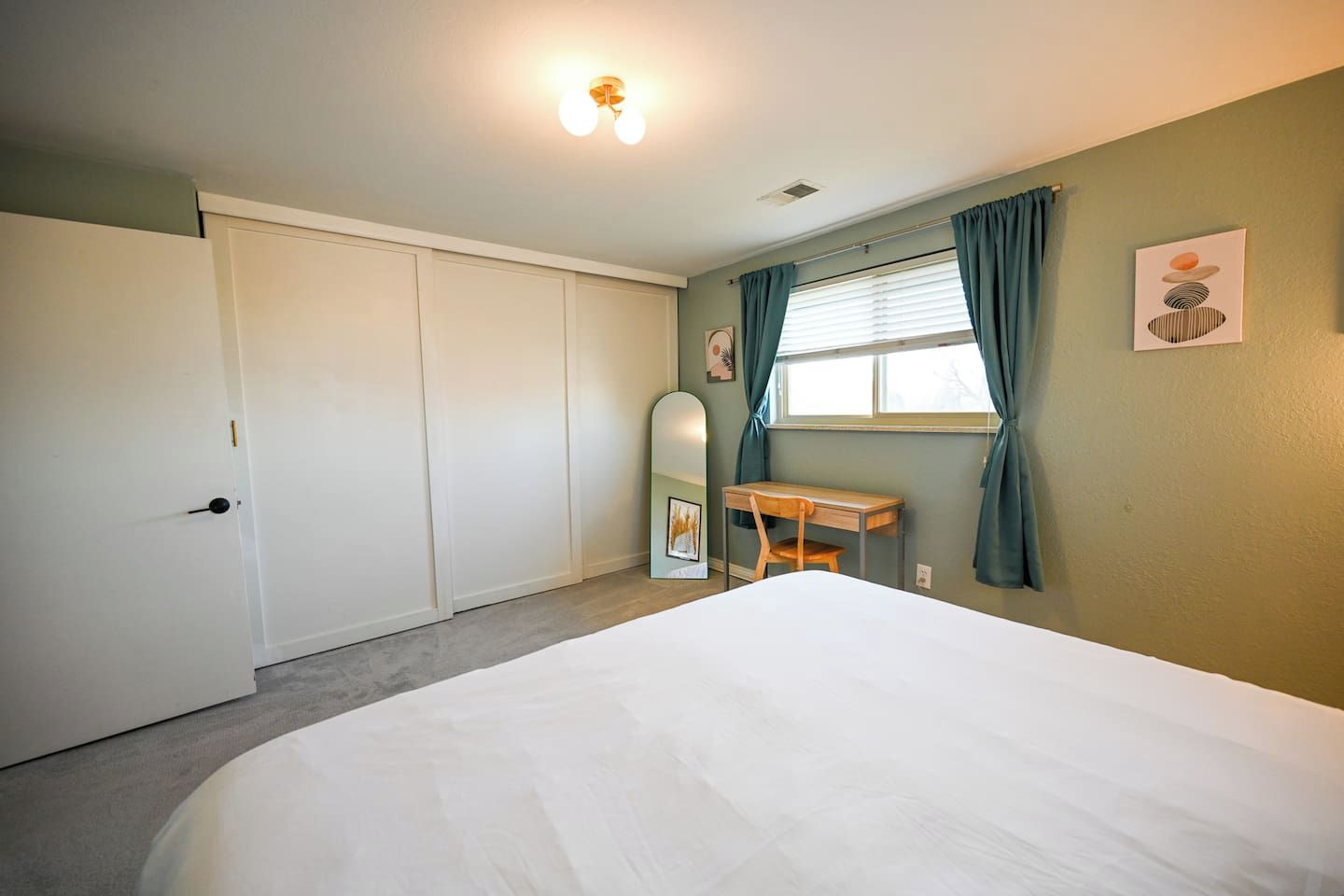 A bedroom with a bed, a desk, a chair, a mirror, and a window with curtains.