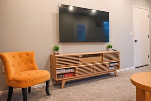 Una habitación con una televisión de pantalla plana montada en la pared, un mueble de madera con espacio de almacenamiento, un sillón naranja con capitoné y una mesita pequeña.