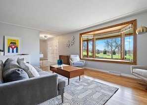 Ein modernes Wohnzimmer mit einer Couch, einem Couchtisch und Blick auf einen Park durch große Fenster.