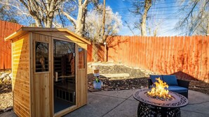 Un cobertizo de madera con puerta de vidrio, un pozo de fuego con llamas y una silla azul en un patio trasero con cerco de madera.