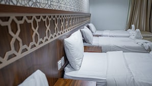 A hotel room with a row of beds, wooden headboard with a geometric pattern, and a wooden nightstand with a remote control. - Saraya Al Misk Hotel (Makkah)