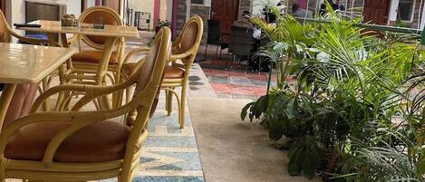 A courtyard with outdoor seating, including wicker chairs and tables, surrounded by potted plants and a tiled floor.