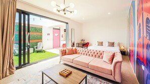 Una habitación de hotel moderna con un amplio ventanal corredero que da a un patio, un sofá rosa, una mesa de centro de madera y una cama con una mesita de noche.