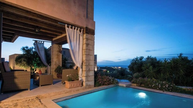 A private pool area with a stone pool, outdoor seating, and a view of the cityscape at dusk. - Pantanassa Villas - Erato with Sea View (Agia Marina)