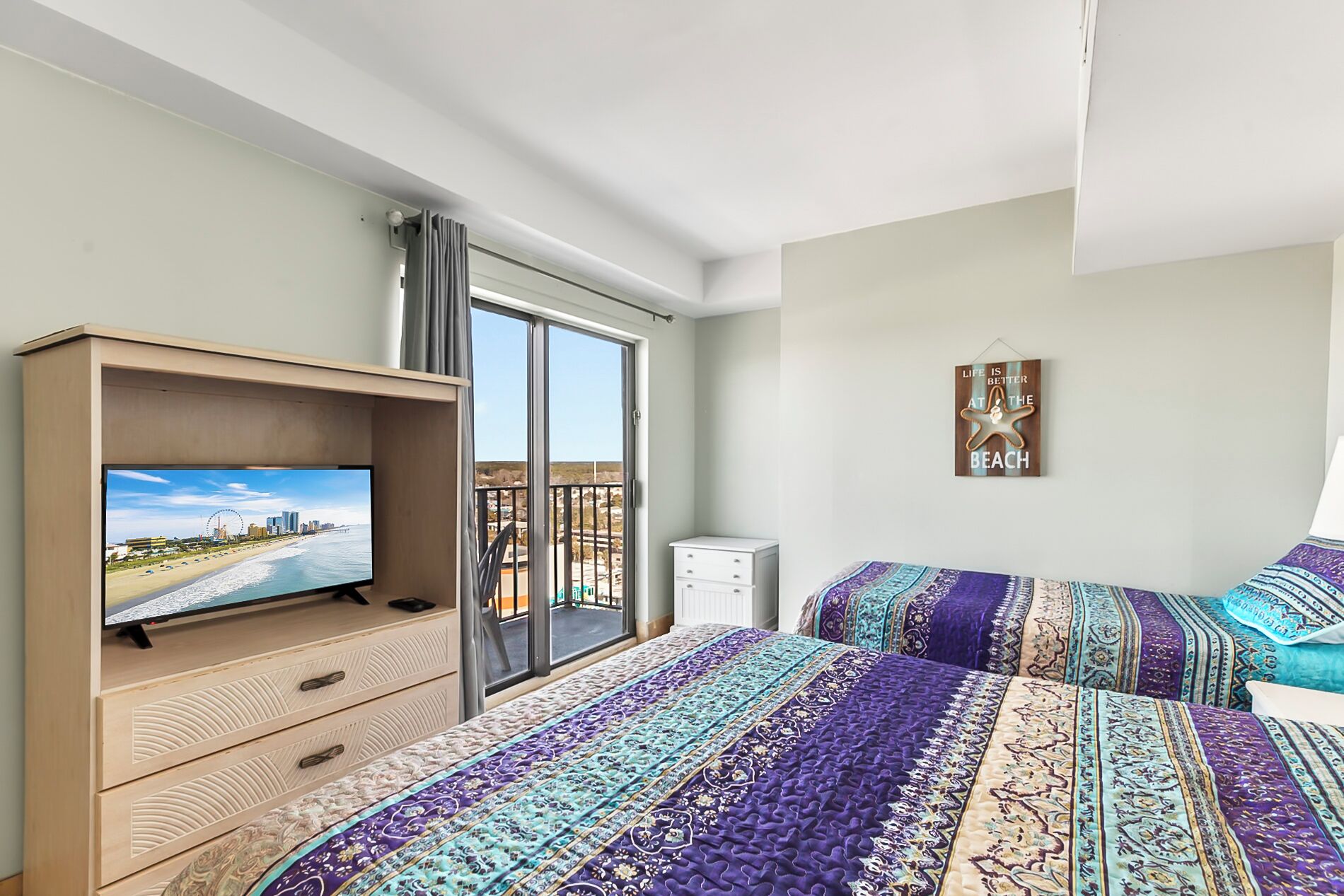 Una habitación de hotel con televisor en un mueble de madera, un balcón con vistas a la playa y cubrecamas estampados.
