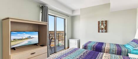 Una habitación de hotel con televisor en un mueble de madera, un balcón con vistas a la playa y cubrecamas estampados.