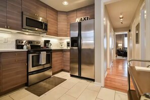 A modern kitchen with stainless steel appliances, wooden cabinets, and a visible 'TAXI' sign on the refrigerator. - 2 Bedroom Duplex Near Food, Shops, & KU Hospital 6 (Kansas City)