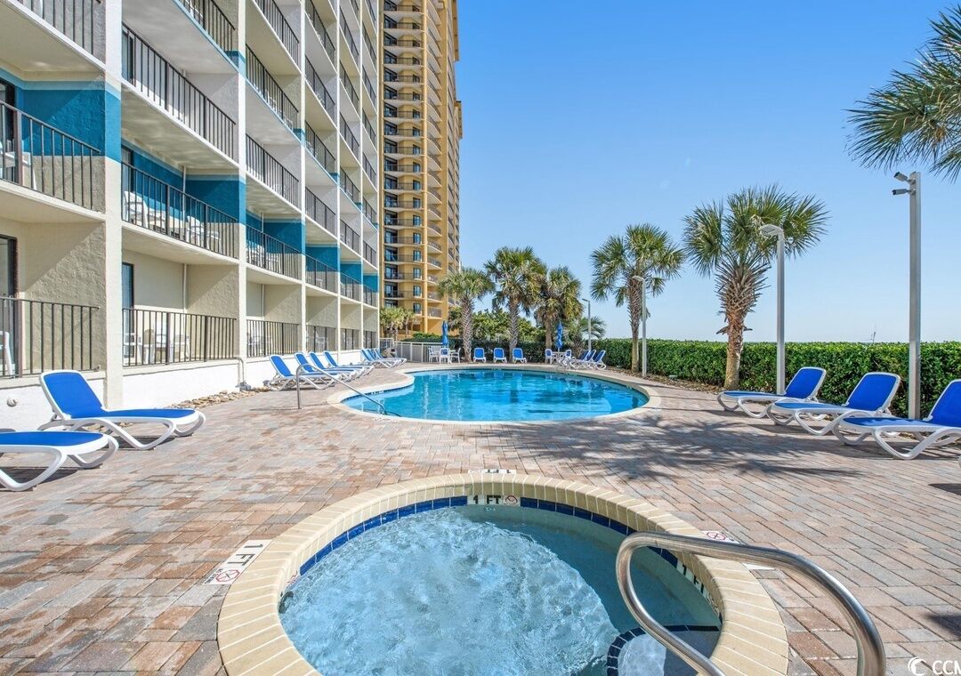 Zona de piscina hotelera con múltiples sillones, un jacuzzi y un edificio alto al fondo.