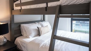 Una cama litera con ropa de cama blanca, una lámpara de noche y una ventana con vistas a edificios.