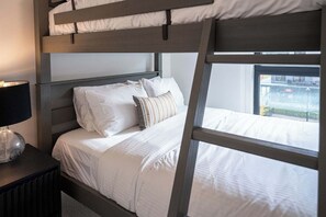 Una cama litera con ropa de cama blanca, una lámpara de noche y una ventana con vistas a edificios.